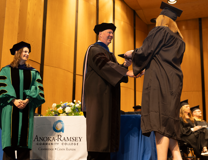 President Hanson hands female graduate her diploma at Cambridge Campus graduation as Vice President looks on