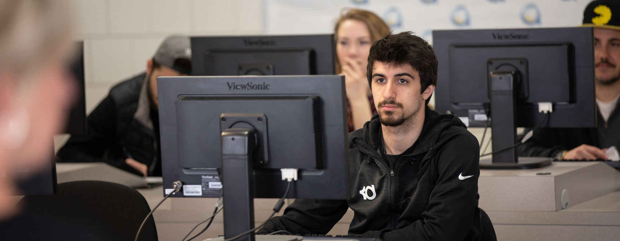 A group of students seated at rows of desktop computers in a campus computer lab, working on assignments during a technology‑focused business class.