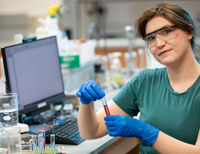 Student in lab with blue gloves and goggles holding test tube with red liquid.