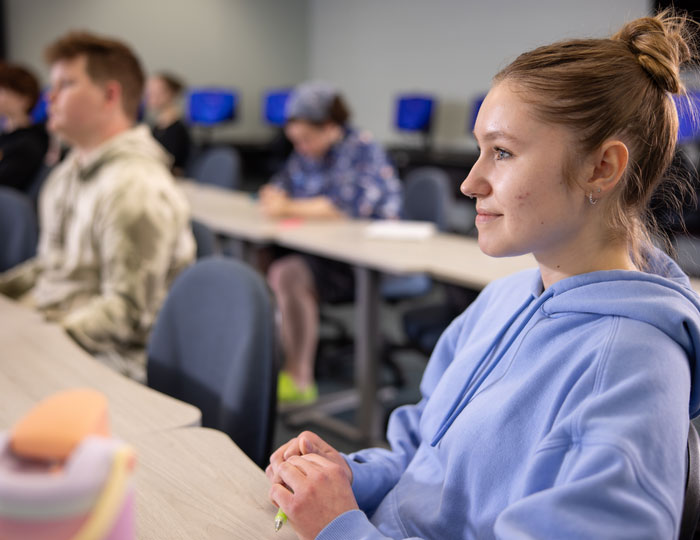 Person sitting at a desk in a computer classroom with other students in background.