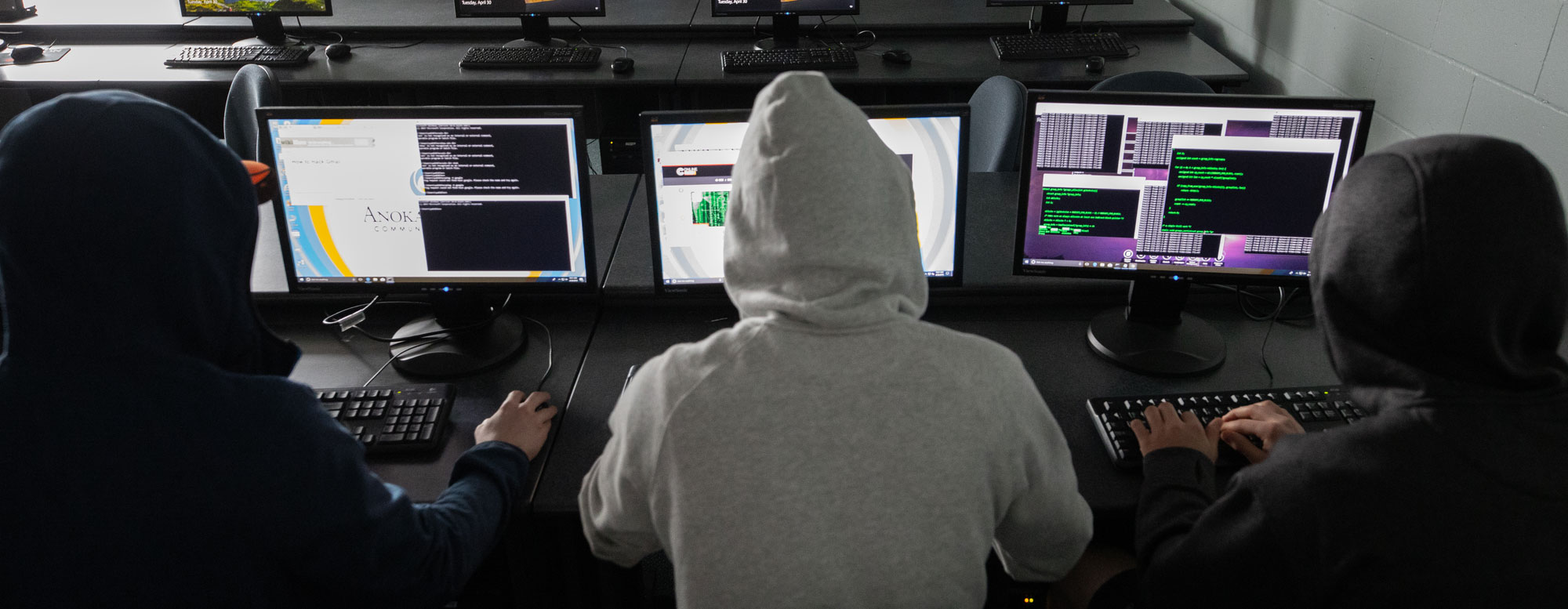 Three people at computer workstations with multiple monitors showing code and technical interfaces.