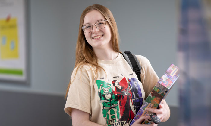 Student standing in a campus hallway holding a boxed project while wearing a graphic T‑shirt and backpack.