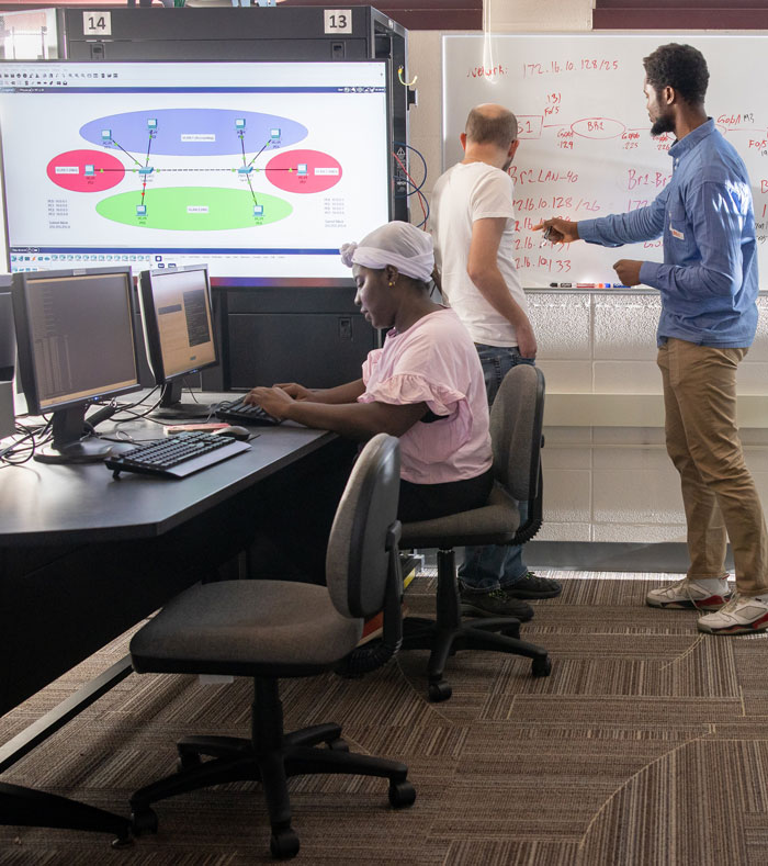 Students working in a computer lab as two classmates discuss a diagram on a whiteboard.