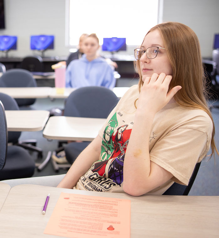 Student seated in a classroom, looking forward thoughtfully with class materials on desk.