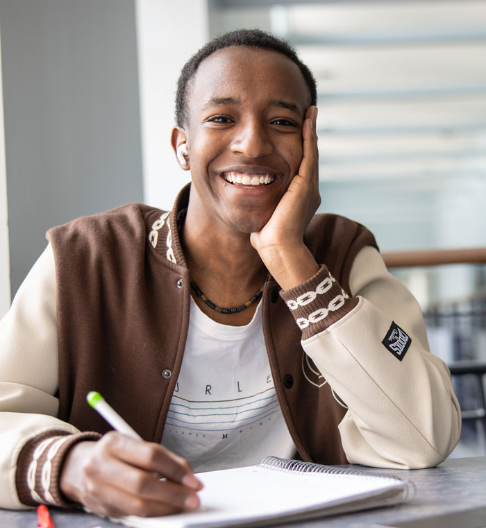 Smiling student seated at a table, resting head on hand while writing in a notebook.