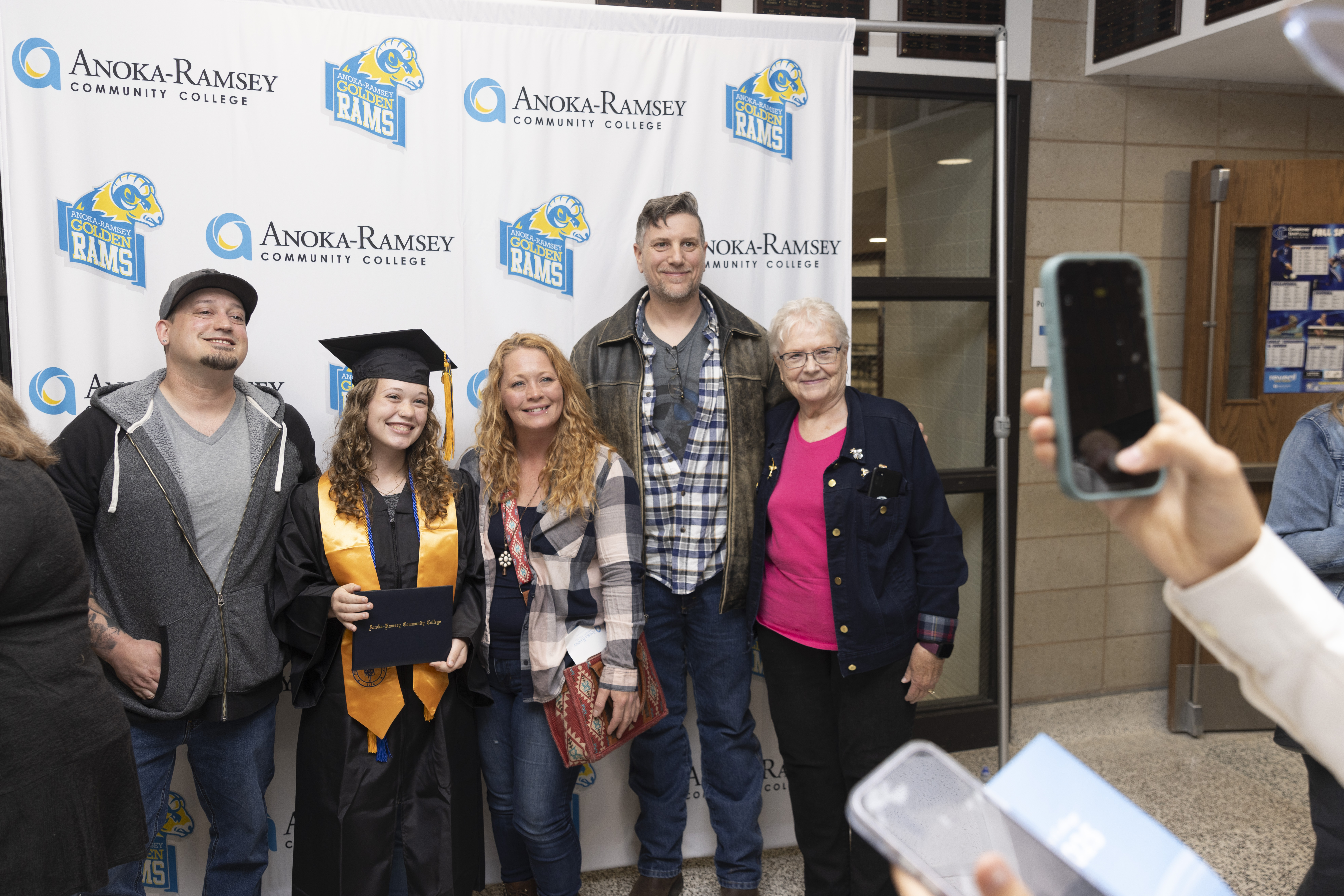 Five individuals, one in graduation robe, posing for photo in front of Anoka-Ramsey photo backdrop