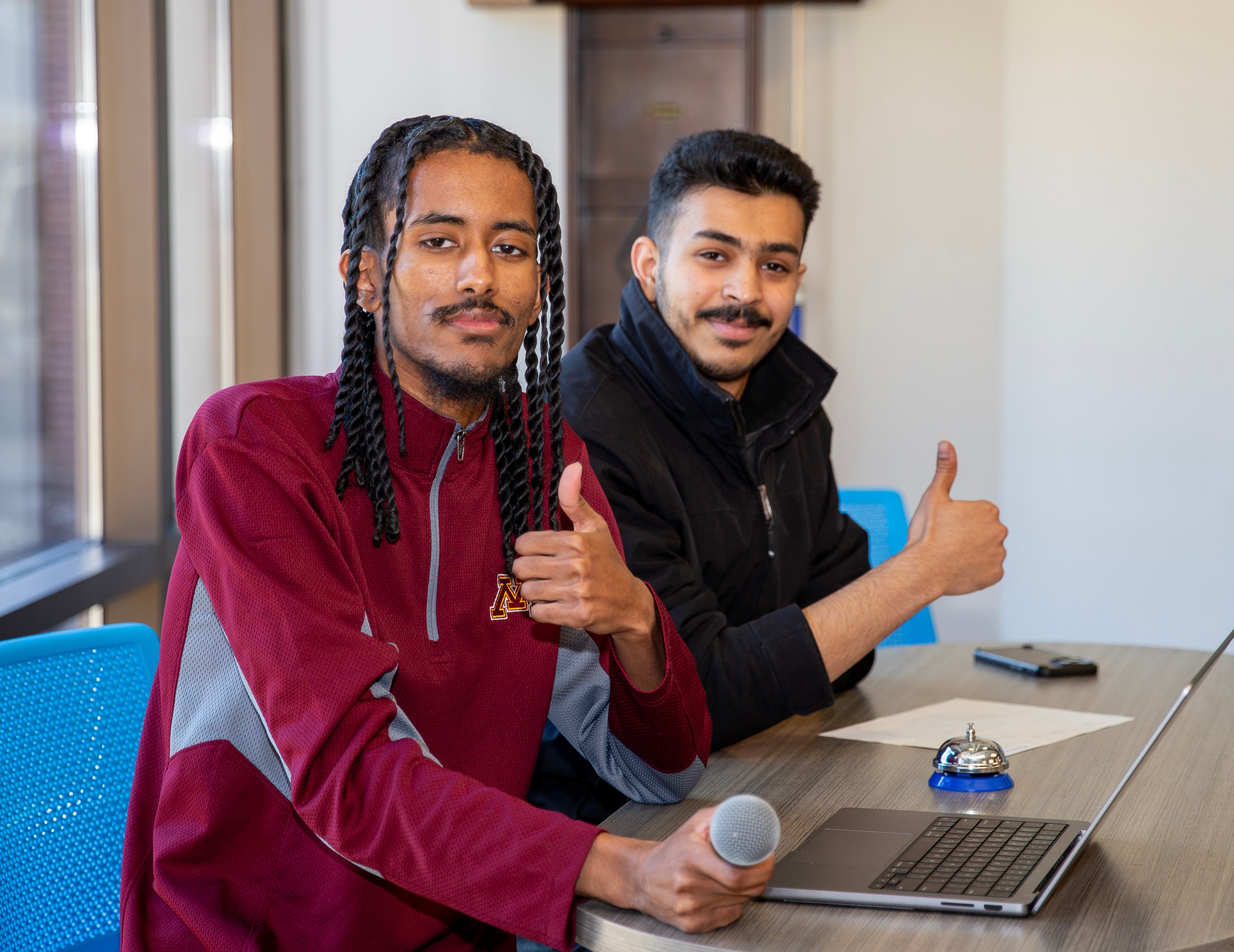two students sitting down at a table looking at the camera, both with their thumbs up