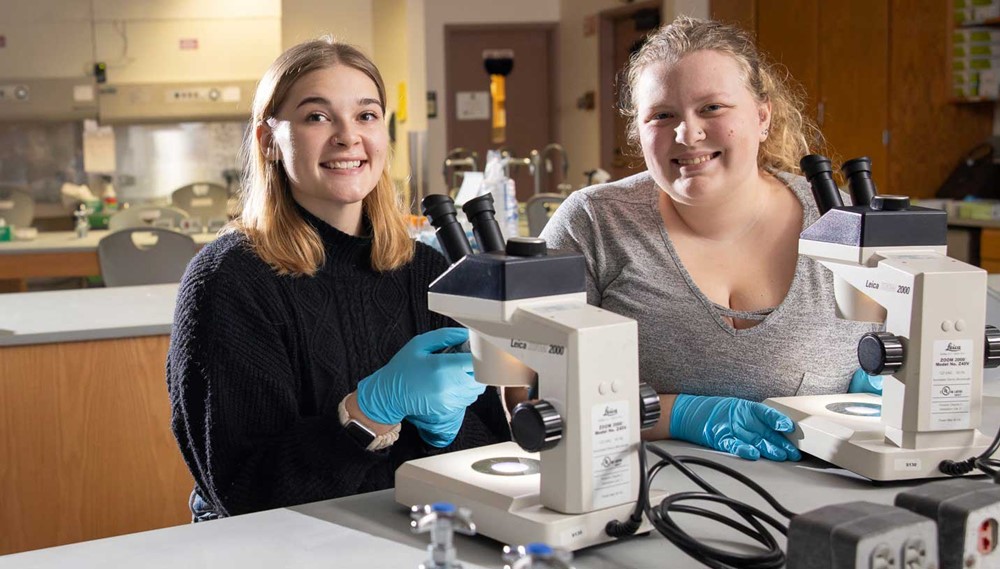 Two individuals in a lab setting, looking up from microscopes at camera.