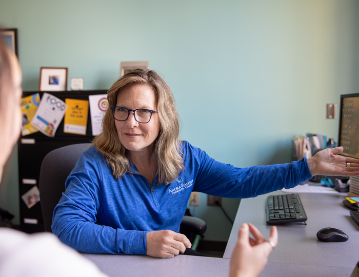Individual pointing at computer screen while speaking to another person in the image's foreground.